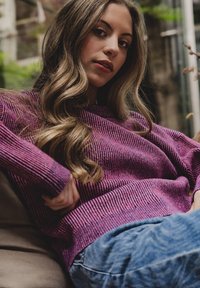 Trui in een gestructureerde donkerroze stof met lange mouwen, ontspannen pasvorm, gecombineerd met blauwe geweven jeans. Zachte achtergrond met groenvoorzieningen.