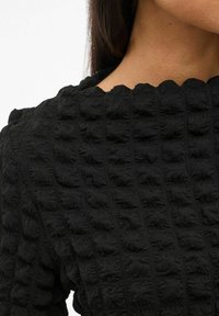 Närbild av en person som bär en svart topp i texturerad tyg med upphöjda fyrkantiga mönster och en scallop-neckline.