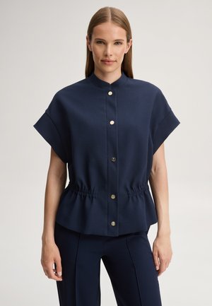 Donna che indossa una camicetta blu navy a maniche corte con bottoni dorati e vita elasticizzata, abbinata a pantaloni blu navy coordinati, in piedi davanti a uno sfondo neutro.