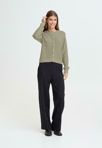 Grön stickad cardigan med rund halsringning, knäppning och ribbade muddarna, kombinerad med svarta vida byxor och svarta skor.