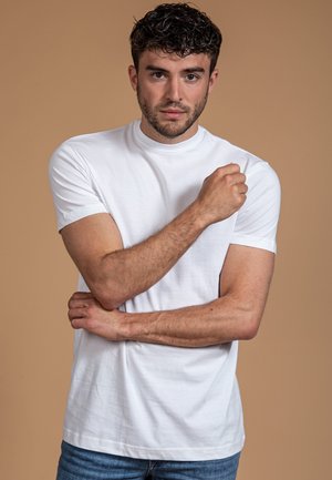 Witte T-shirt met korte mouwen in een losse pasvorm, gemaakt van soepel katoen, gecombineerd met blauwe denim jeans. Model staat met gekruiste armen.
