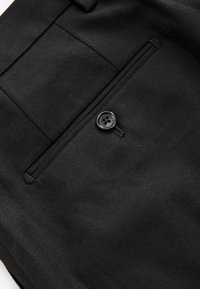Gros plan sur un tissu noir montrant une poche ajustée avec un seul bouton noir et une boutonnière cousue.