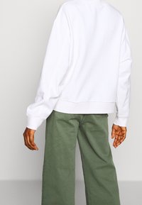 Witte sweatshirt met uitgevallen schouders en ribbelmanchetten, gecombineerd met lichtgroene wijdlopende broeken, gezien van achteren. Zacht, casual materiaal.