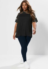 Czarna koszulka z przezroczystymi, kwiatowymi rękawami, zestawiona z ciemnoniebieskimi legginsami i białymi trampkami. Gładka tekstura, luźny krój i zwiewny design.