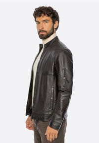 Mężczyzna z kręconymi włosami i brodą, ubrany w czarną skórzaną kurtkę na białym golfie, stojący bokiem na jednolitym tle.