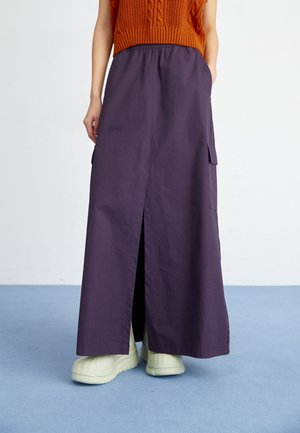 Mannequin portant une longue jupe violette foncée avec une fente à l'avant, associée à des baskets épaisses de couleur crème claire et un haut sans manches en maille orange.