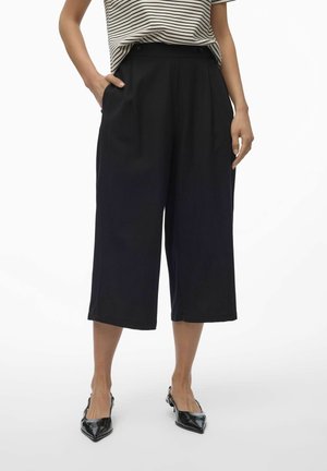 Vrouw draagt zwarte wijde cropped broek, gestreept top met korte mouwen en zwarte slingback schoenen met puntige neus, staand met handen in de zakken.