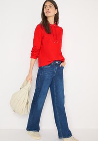 Czerwona bluza z kapturem i sznurkami, zestawiona z szerokimi niebieskimi dżinsami o szerokich nogawkach. Modelka trzyma jasnobeżową torebkę.