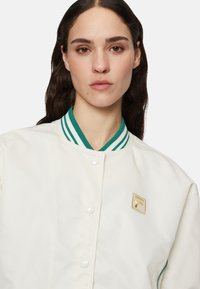 Krämfärgad bomberjacka med grönrandig krage, tryckknappsstängning och en guldbroderad logotyp på bröstet.