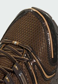 Ruskea urheilukenkä, jossa on teksturoitu verkkopäällinen, kiiltävät korostukset ja kuvioitu nauhamalli. Sivulla huomattavia heijastavia yksityiskohtia.