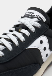 Black leather sneakers with white laces, featuring a woven texture, a distinct white logo and accents, and perforated detailing on the side.