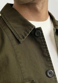 Giacca verde oliva con colletto a punta, due bottoni neri e una texture liscia, indossata sopra una camicia bianca.