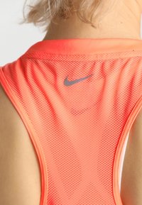 Ein korallenfarbenes Nike-Athletik-Top mit Netzstruktur, das ein tone-in-tone silbernes Nike-Logo im Bereich des Nackenrückens aufweist.