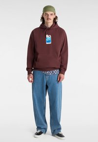 Vinröd hoodie med en känguruficka, i kombination med stora ljusblå jeans. En extra blå rutiga skjorta syns under hoodien.