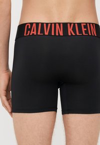 Boxeri negri din material elastic, cu un talie lată pe care este imprimat în litere roșii îndrăznețe "CALVIN KLEIN".