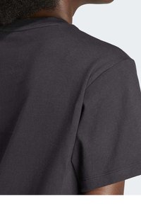 Ung person med mörk hud och lockigt hår, klädd i en enkel svart t-shirt med kort ärm, sedd bakifrån med huvudet lätt vänd.