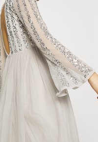 Une robe gris clair présente de longues manches transparentes ornées de sequins argentés et un dos dénudé, mettant en valeur une jupe fluide et texturée.