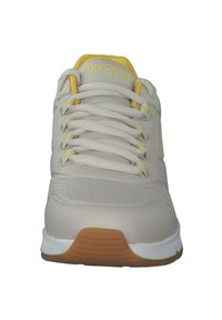 Vaaleanbeige urheilukengät, joissa on kuvioitu nahkapäällinen, keltaisia yksityiskohtia ja valkoinen kumipohja. Kengissä on pehmustettu kieli ja kuusi nyörireikää.