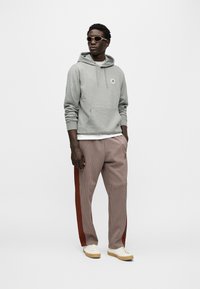 Γκρι φούτερ με κουκούλα και μπροστινή τσέπη με λογότυπο, συνδυασμένο με καρό παντελόνι με κόκκινες πλευρικές ρίγες. Ολοκληρώνεται με μπεζ αθλητικά παπούτσια και γυαλιά ηλίου.