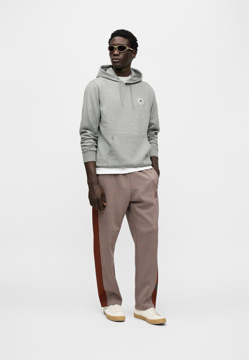 Grå hættetrøje med forlomme og logo, kombineret med ternede bukser med røde sidelister. Fuldført med beige sneakers og solbriller.