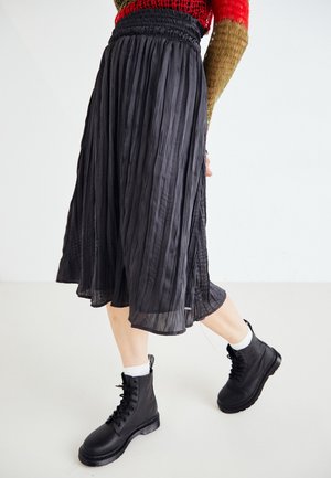 Jupe midi plissée noire avec une taille élastique, un matériau drapant et un éclat subtil, associée à des bottes de combat noires et des chaussettes blanches.