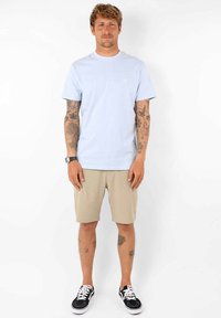 T-shirt en coton bleu clair avec un col rond, associé à un short beige et des baskets noires avec des lacets blancs. Tatouages visibles sur les bras.