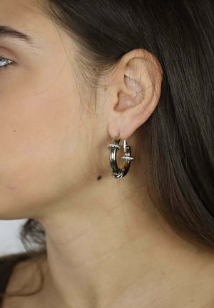 Silberne Creolen mit glatter Oberfläche, dekorativen Akzenten und einem Verschluss, die von einem durchstochenen Ohr hängen.