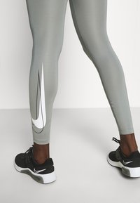 Szare legginsy z białym logo Nike, charakteryzujące się gładką, elastyczną teksturą. W zestawieniu z czarnymi sneakerami, prezentując nowoczesną estetykę sportową.