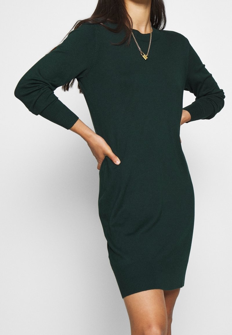 Femme portant une robe en maille vert foncé à manches longues avec un collier chaîne doré, posant les mains sur les hanches devant un fond blanc.