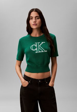 Frau mit langen dunklen Haaren, die einen grünen, kurz geschnittenen Pullover mit kurzen Ärmeln und "cK"-Logo sowie schwarze Hose trägt, Hände in den Taschen, neutraler Hintergrund.