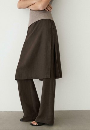 Frau trägt dunkelbraunen, mehrlagigen Rock über passenden weiten Hosen mit schwarzen offenen Sandalen, die Arme verschränkt, vor schlichtem Wandhintergrund.