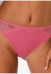 Rožnate ženske spodnje hlačke z čipko, nošene nizko na pasu, z vidno kožo in delnim pogledom na roko, ki počiva na stegnu.