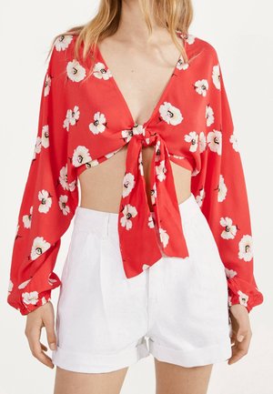 Nainen, jolla on päällään punainen, kukallinen, pitkähihaisen lyhyt pusero, joka on solmittu edestä, yhdistettynä korkeavyötäröisiin valkoisiin shortseihin, seisoo yksivärisen taustan edessä.