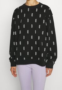 Felpa nera con il logo bianco "HUGO" ripetuto, collo rotondo, polsini e fondo a costine. Tessuto elastico con vestibilità rilassata, abbinata a pantaloni lavanda.