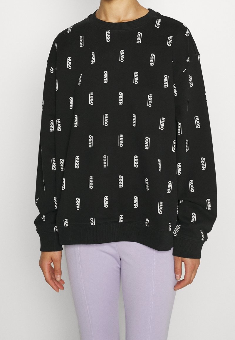 Felpa nera con il logo bianco "HUGO" ripetuto, collo rotondo, polsini e fondo a costine. Tessuto elastico con vestibilità rilassata, abbinata a pantaloni lavanda.