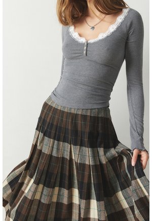 Žena v šedém topu s dlouhým rukávem, krajkovým výstřihem a knoflíky, v kombinaci s plisovanou vícenásobně barevnou kostkovanou sukní, držící okraj sukně.
