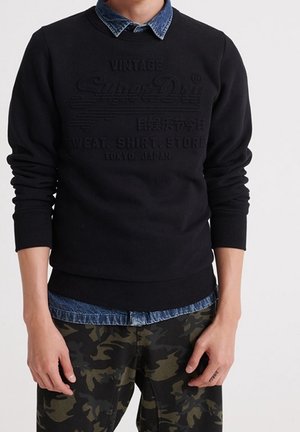 Hombre vestido con sudadera negra sobre una camisa de mezclilla azul y pantalones de camuflaje, de pie con las manos relajadas a los lados.