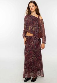 Mujer joven con una blusa de hombros al aire en color burdeos con estampado paisley y una falda larga a juego, llevando un bolso de hombro y usando unos zapatos planos negros.