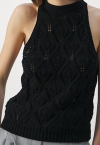 Musta neuleinen halter-top, jossa on timantti- ja aalto-kuviointi, ribatulla helman ja avoimella kudonnalla, suunniteltu istuvalle siluetille.