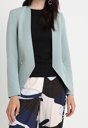 Kvinde iført en lyseblå blazer over en sort ribbet top og løse bukser med abstrakte mønstre i marineblå, sort og hvid.