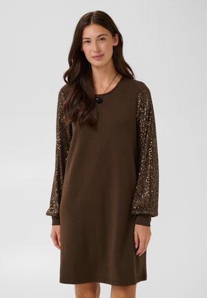 Mujer con un vestido marrón con mangas largas brillantes cubiertas de lentejuelas y un collar negro con colgante, de pie frente a un fondo claro y liso.
