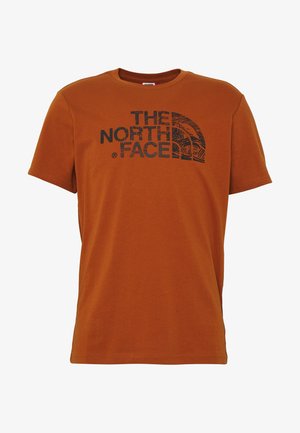 Rdzawy t-shirt z krótkim rękawem z czarnym logo "The North Face" i grafiką półkopuły na środku klatki piersiowej.
