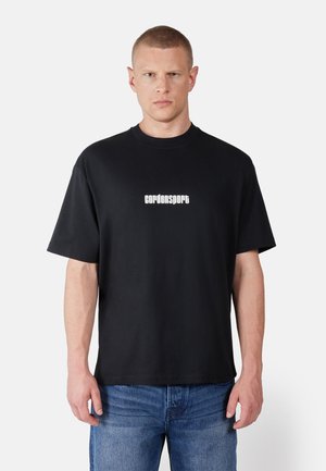 Man with short hair wearing black Cordonsport t-shirt and blue jeans, standing against plain light background.