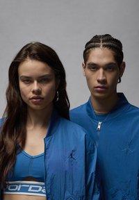 Cazadoras de rendimiento azules con frentes con cremallera, que presentan acentos del logo de Air Jordan. La tela es suave con una textura ligeramente reflectante.