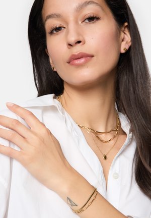Goldschmuck, einschließlich geschichteter Halsketten, eines Anhängers und eines Armbands, kombiniert mit einem weißen Hemd. Geschmeidige Haut und lange dunkle Haare sind sichtbar.