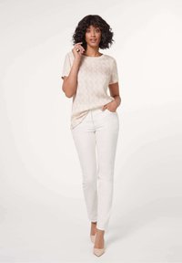 Lichtbeige T-shirt met korte mouwen en een subtiel logo patroon, gecombineerd met witte strakke broek en nude hoge hakken, staand tegen een neutrale achtergrond.