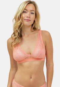 Bralette en dentelle corail clair avec un design triangulaire, des bords festonnés et un petit accent central. Texture douce sur une bande lisse.