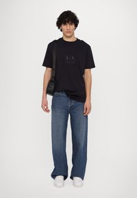 Camiseta de algodón azul oscuro con logo en relieve, combinada con vaqueros de pierna ancha azul y zapatillas blancas. Pequeño bolso negro bandolera con diseño texturizado.