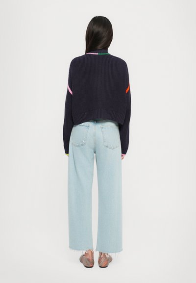 Marineblauwe ribbelgehaakte cropped sweater met kleurrijke accentstrepen, gecombineerd met lichtblauwe wijde spijkerbroek en gemusterde instappers.