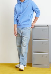 Ljusblå huvtröja med framficka och vitt logo, kombinerad med ljusblå jeans och vita sneakers med beige detaljer.